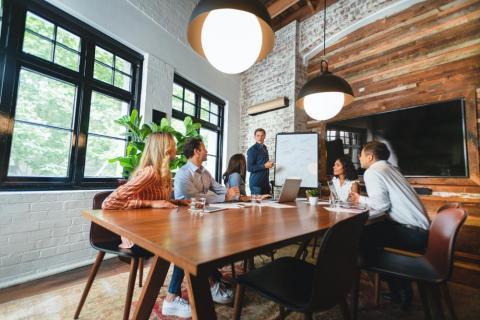 Three women and two men sitting at a conference table listening to a man presenting at a whiteboard