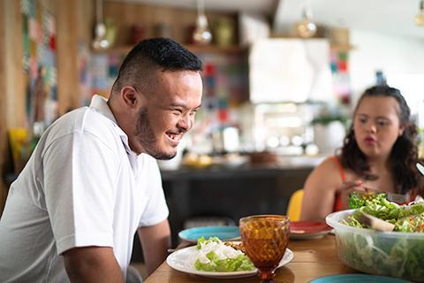 Man and woman sitting at a dining table. Man is smiling and there is a bowl of salad between them.
