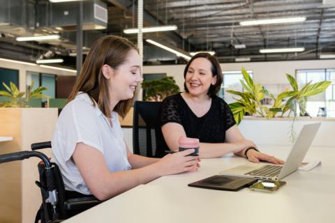 Woman sitting in a wheelchair at a desk with another woman sitting next to her. There is a laptop on the desk and the woman using the wheelchair holds a reusable coffee cup. The two women are having a conversation.