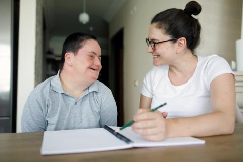 A woman wearing glasses and a man with down syndrome sitting at a table smiling at each other. There is a notebook on the table and the woman is about to write in the notebook with a pencil.
